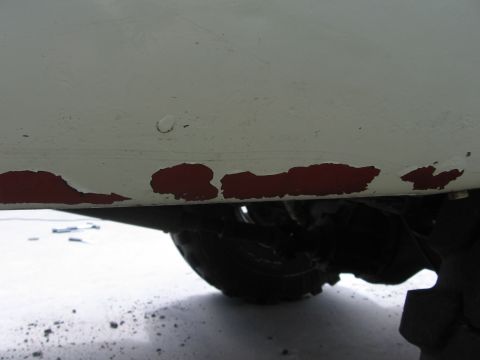 1979 Ford F-150 - Detail of a white truck's lower body panel reveals chipped red paint, with a tire and suspension visible underneath.