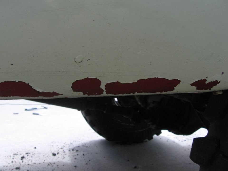 White truck lower body panel with chipped red paint, showing tire and suspension components