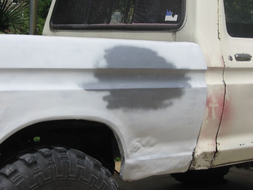 Side view of a white truck's rear fender and bed, showing body filler and primer. Tire visible