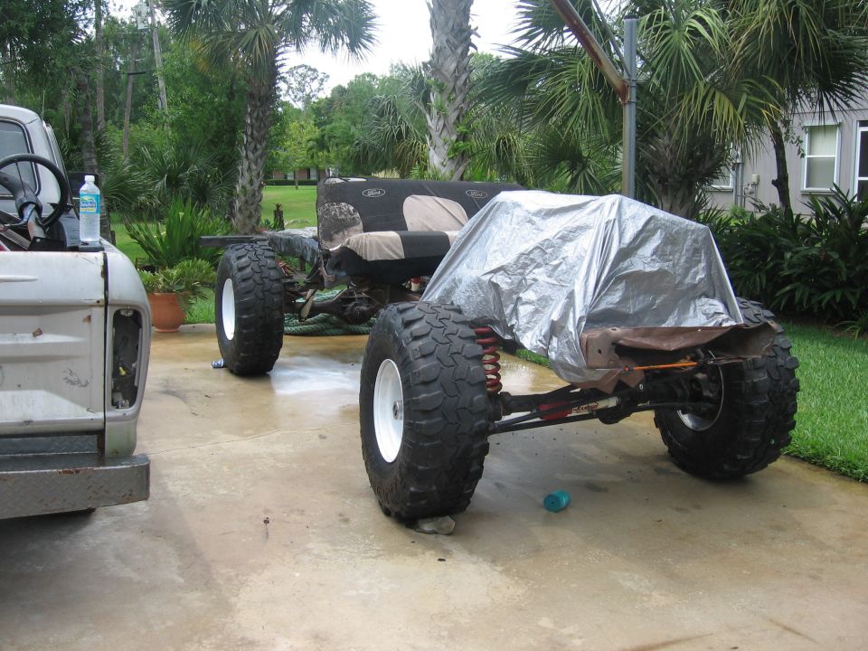 Two custom off-road chassis in a driveway, one partially covered with a tarp. A bottle of water sits on the fender of the truck on the left