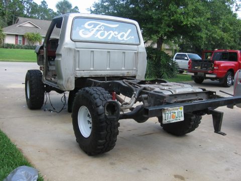 1979 Ford F-150 - A bare Ford truck chassis with a cab, modified with large off-road tires, sits in a driveway with other vehicles in the background.
