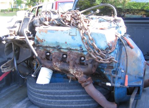 1979 Ford F-150 - A heavily rusted engine block, painted blue, is secured with chains and sits on a tire in the back of a pickup truck.