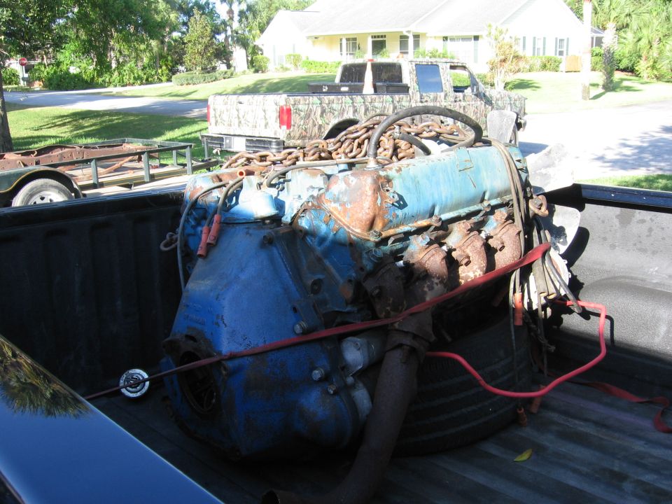 Blue engine with rusty parts and chains secured in a truck bed. A trailer is visible behind