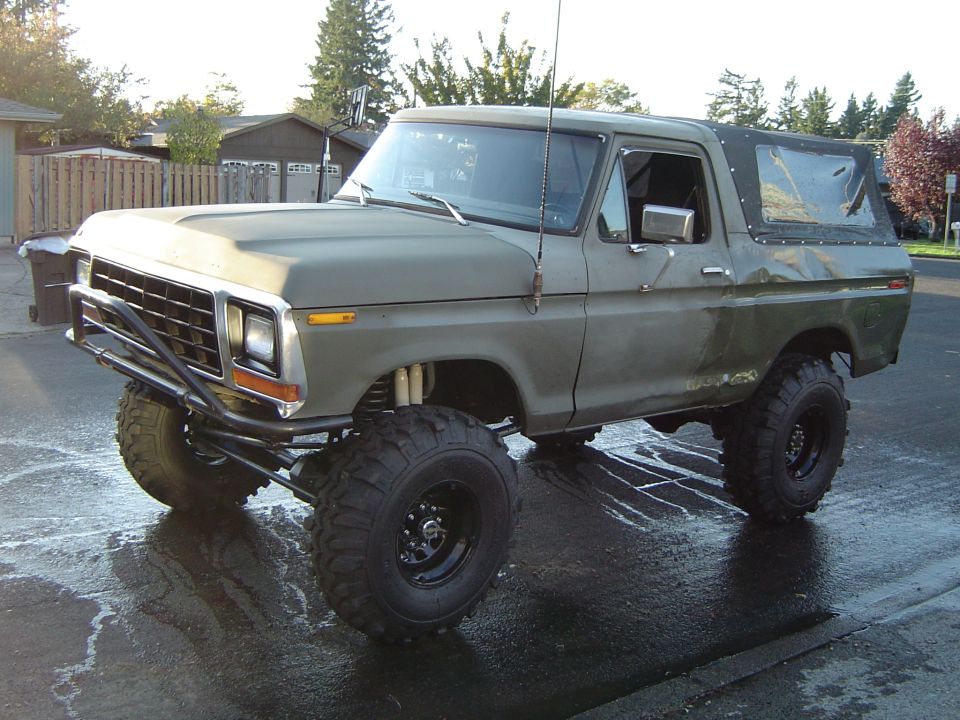 Olive green 1978 Ford Bronco with lifted suspension and large tires parked on a wet asphalt surface