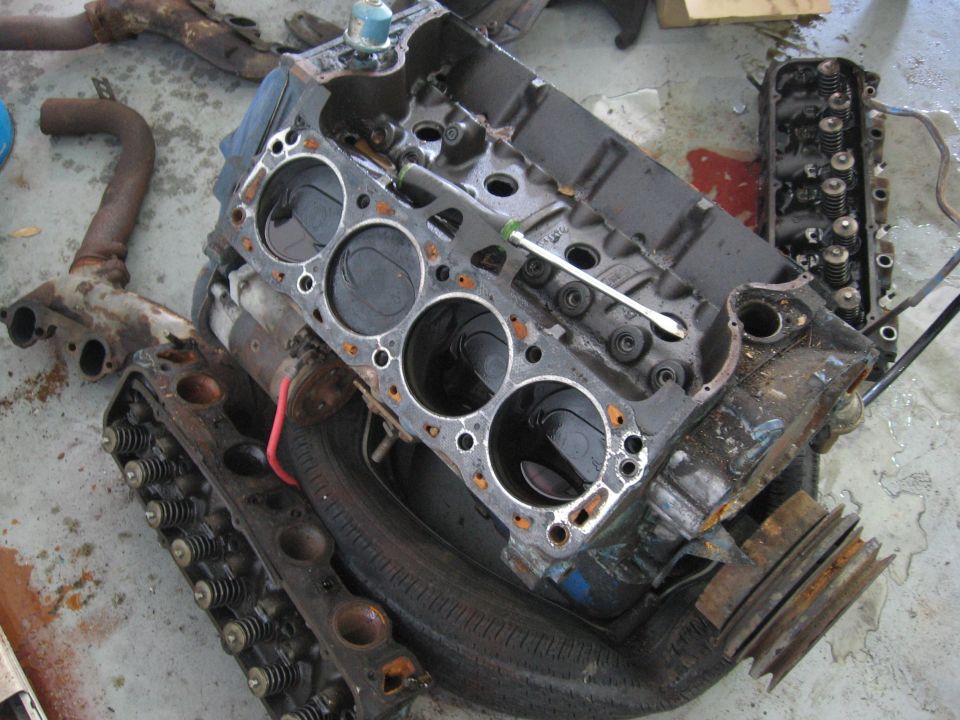Disassembled V8 engine block and cylinder heads on a concrete floor. A screwdriver rests on the block