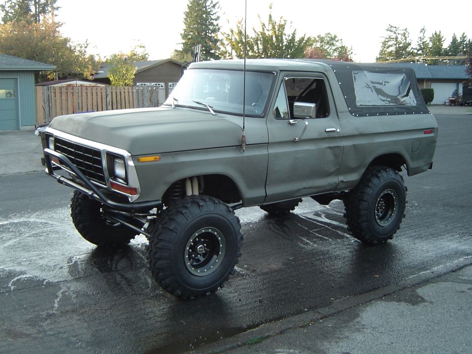 Olive-drab lifted full-size Ford Bronco with soft top and big bar-tread tires on a wet driveway