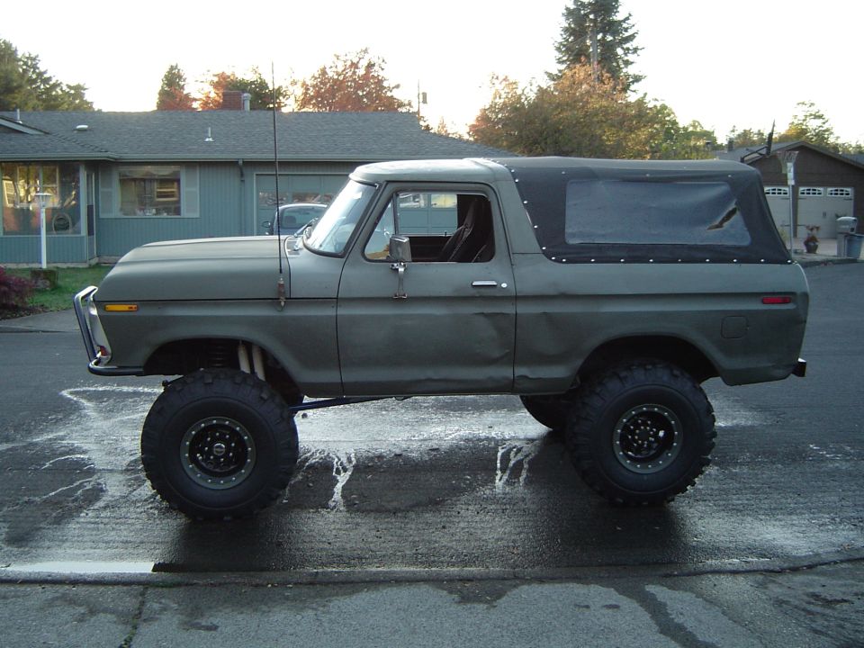 Flat green lifted Ford Bronco with black soft top on mud tires, side view on wet street