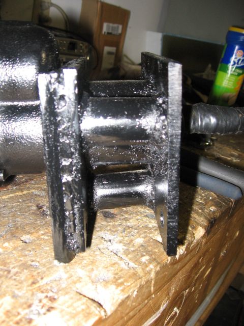 1979 Ford F-150 - Close-up of a heavy black-painted cast bracket standing on a chewed-up wooden workbench, its two flanges and ribbed web showing fresh grind…