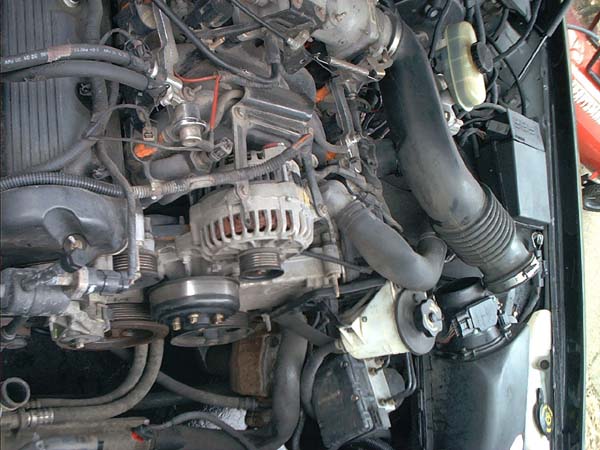 Engine bay overhead view showing alternator, serpentine belt and black corrugated air intake tube