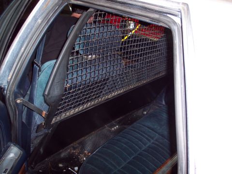 1991 Ford LTD Crown Victoria - interior of a car shows a metal wire-mesh prisoner cage partition mounted behind worn blue cloth front seats.