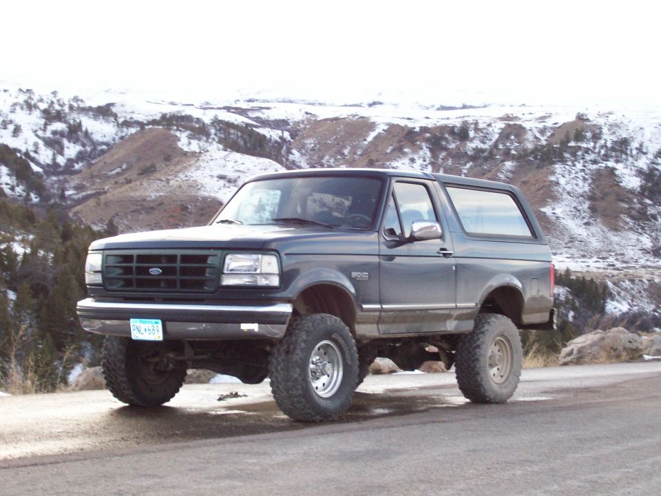 Dark gray 1994 Ford Bronco with lifted suspension parked on a dirt road with snowy hills in the background
