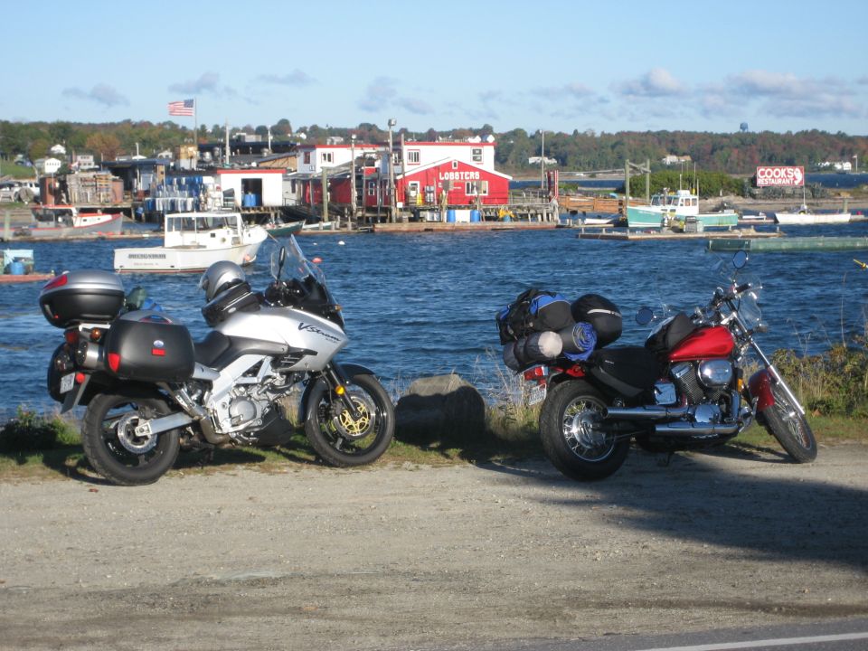 Red motorcycle parked on a gravel path with luggage on the back, facing right. A silver motorcycle is parked to its left