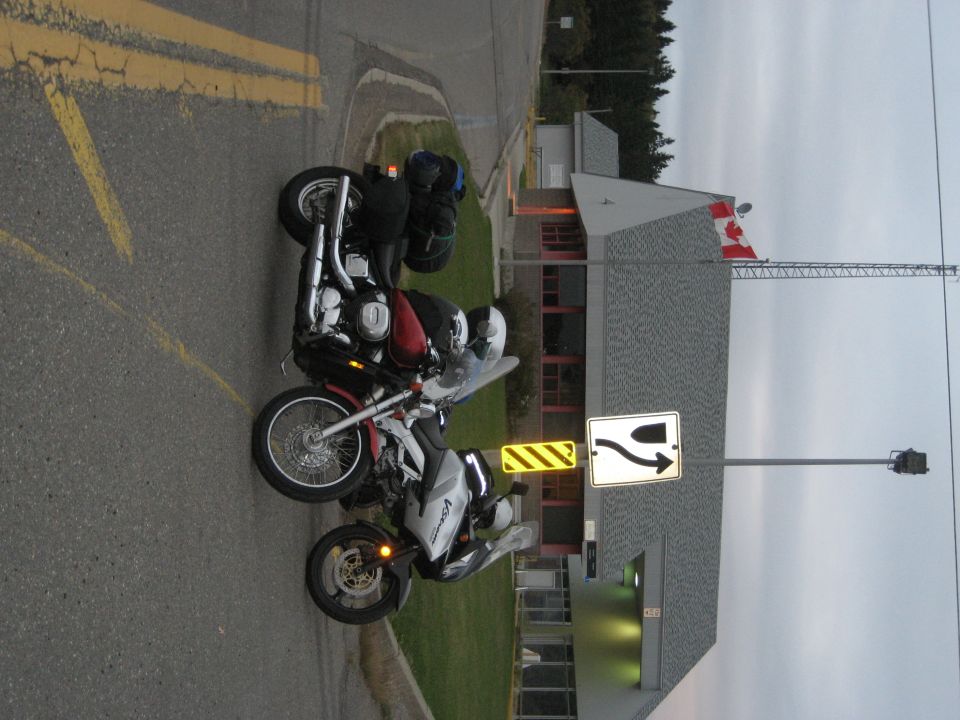 Two motorcycles parked on a street, one red and black, the other silver and black, with a building and Canadian flag in the background
