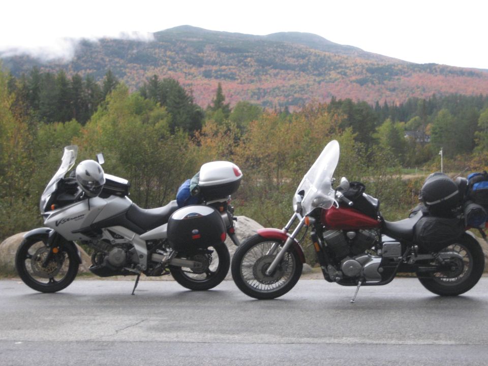 Two touring motorcycles parked on a roadside with autumn foliage and mountains in the background