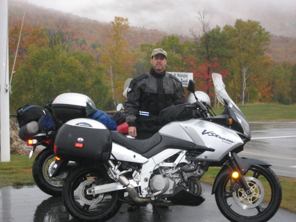 Man in riding gear stands by a silver Suzuki V-Strom motorcycle. Another motorcycle is parked behind it