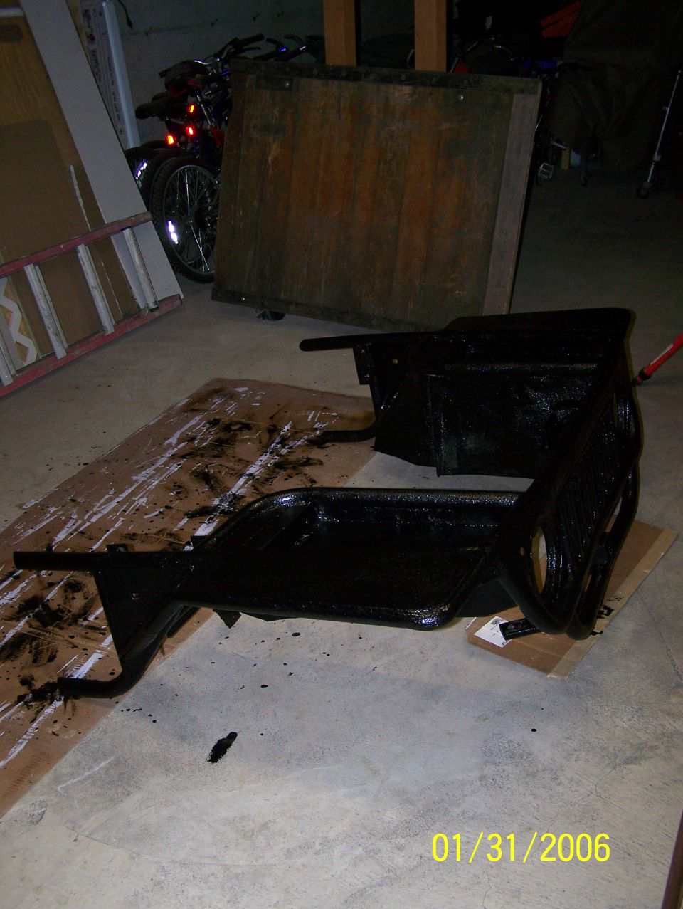 Two freshly black-painted vehicle frame or bracket parts drying on cardboard in a garage