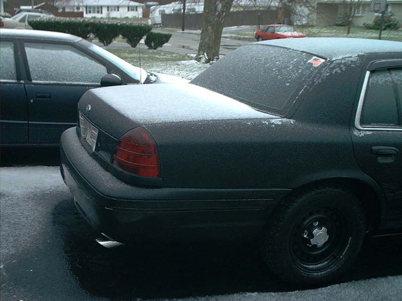 Rear quarter view of a dark sedan covered in snow, parked next to another car. A red car is visible in the background