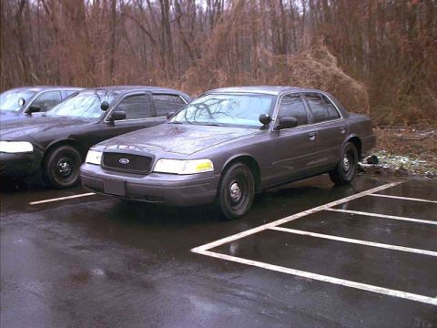 2000 Ford Crown Victoria - Three-quarter front view of a gray Ford Crown Victoria P71 police-package sedan parked on wet, white-striped asphalt, with black steel whee…