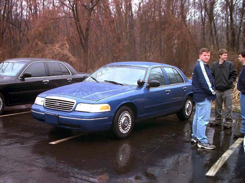 Blue Ford Crown Victoria with steel wheels and hubcaps, 3/4 front, on wet pavement