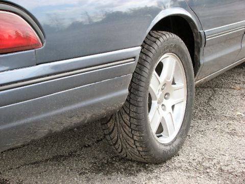 1985 Mercury Grand Marquis - The rear side view of a gray sedan reveals its tire and fender, with a visible SuperMotors watermark.