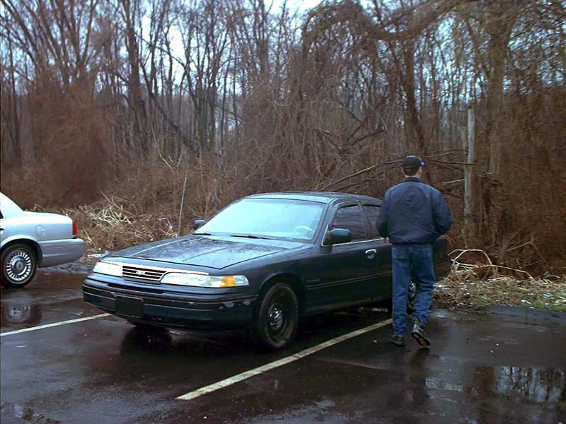Dark blue Ford Crown Victoria parked on wet asphalt, driver standing nearby