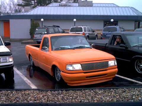 2000 Ford Crown Victoria - An orange Ford Ranger pickup truck is parked in a lot next to other vehicles, with a person visible in the black truck.