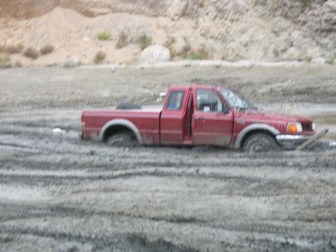 1987 Ford Bronco - A red pickup truck is stuck in thick mud, with its doors open and wheels submerged.