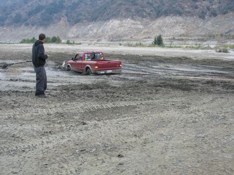 1987 Ford Bronco - A red pickup truck is stuck in thick mud, with a person standing nearby observing the situation.
