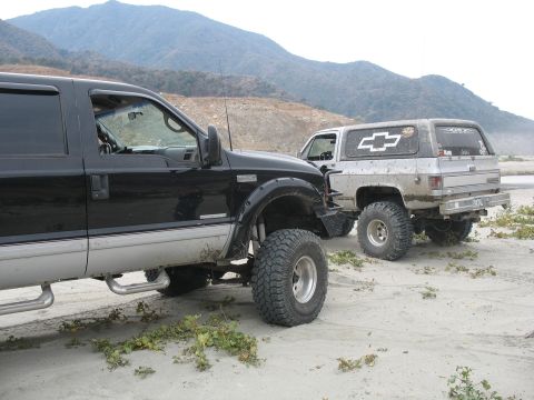 1987 Ford Bronco - Two lifted trucks, a Ford Super Duty and a Chevrolet K5 Blazer, are parked on a sandy area with mountains in the distance.