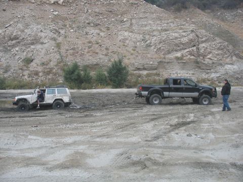 1987 Ford Bronco - Two off-road vehicles are stuck in a muddy area with a rocky hillside in the background.