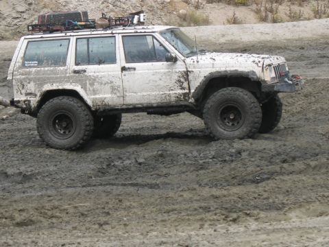1987 Ford Bronco - A white Jeep Cherokee navigates a muddy terrain, equipped with a roof rack and off-road tires.
