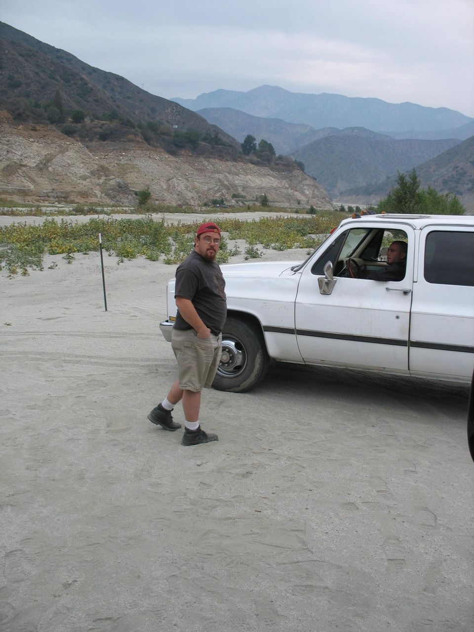 Man in shorts and t-shirt stands next to white SUV; driver visible inside. Mountains and dry brush in background
