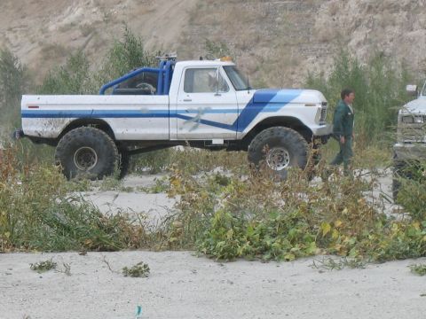 1987 Ford Bronco - A man walks past a modified pickup truck with large tires and a custom roll cage in an outdoor, dusty environment.