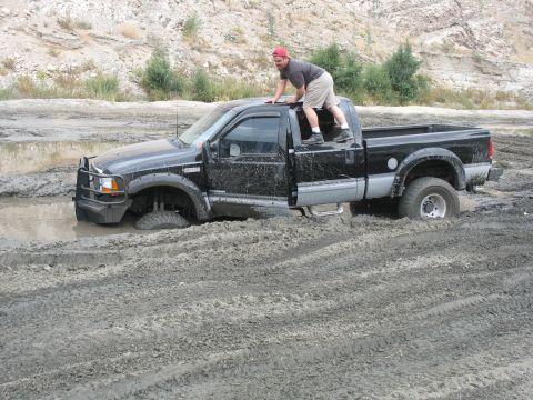 1987 Ford Bronco - A man climbs on top of a black pickup truck that is stuck in a muddy area.