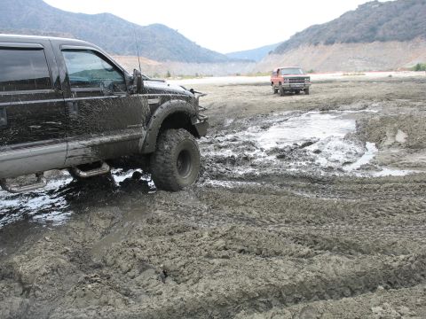 1987 Ford Bronco - Two off-road vehicles navigate a challenging muddy terrain with hills in the background.