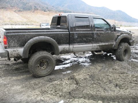 1987 Ford Bronco - A black pickup truck navigates a muddy terrain, with a white SUV visible in the background.