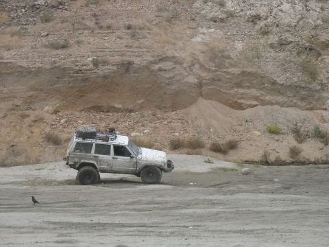 1987 Ford Bronco - A white Jeep Cherokee XJ with a roof rack and spare tire is parked on a dirt road with a rocky hillside in the background.