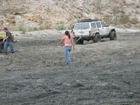 1987 Ford Bronco - A white Jeep Cherokee XJ is stuck in a muddy area, with two people nearby.
