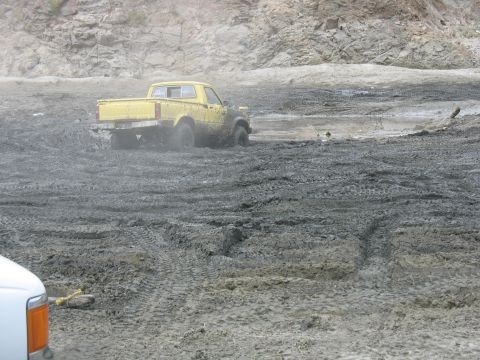 1987 Ford Bronco - A yellow pickup truck is stuck in thick mud, with part of another white vehicle visible in the foreground.