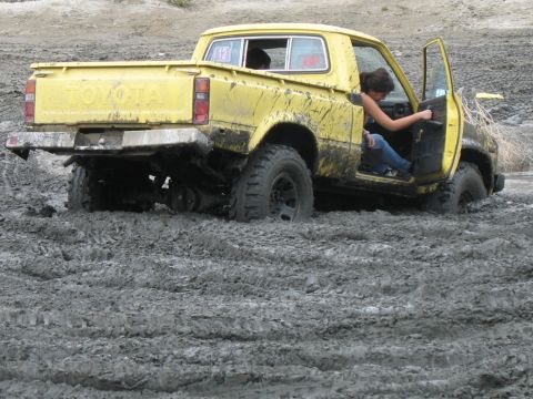 1987 Ford Bronco - Yellow Toyota pickup truck is stuck in thick mud with the driver's door open.