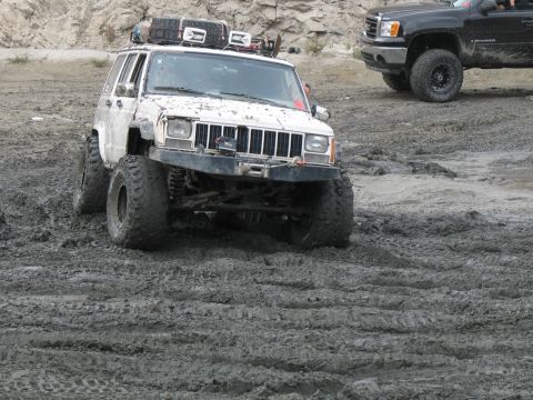 1987 Ford Bronco - A white Jeep Cherokee XJ navigates a muddy terrain, with a black pickup truck visible in the background.
