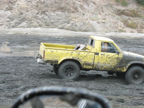 1987 Ford Bronco - A yellow pickup truck navigates a muddy terrain, its body heavily splattered with mud.