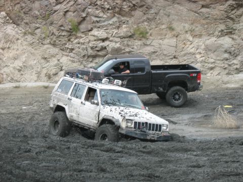 1987 Ford Bronco - Two off-road vehicles navigate a muddy terrain with rocky hills in the background.