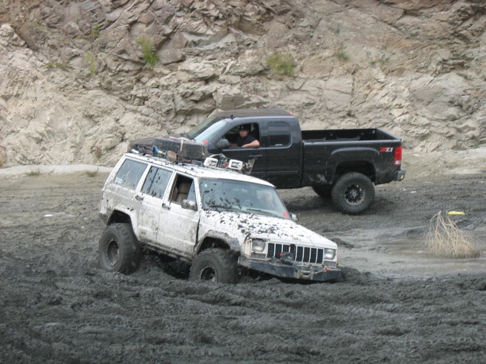 White Jeep Cherokee stuck in mud, front right tire submerged. Black pickup truck behind it