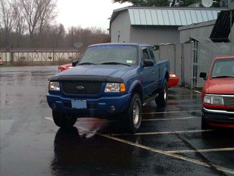 2000 Ford Crown Victoria - A blue Ford Ranger pickup truck is parked in a lot on a wet day, with a red vehicle visible to its right.