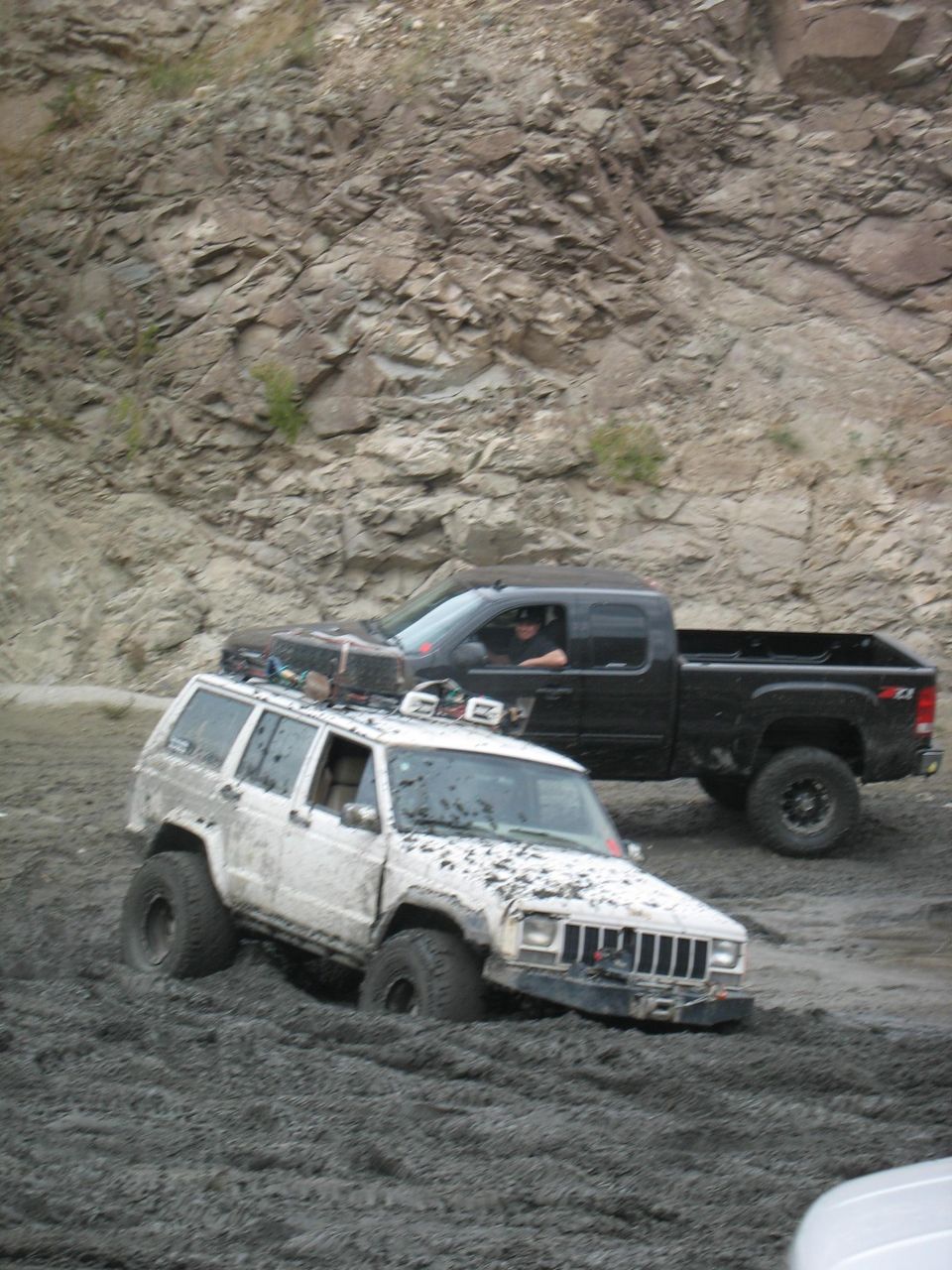 White Jeep Cherokee stuck in mud, black pickup truck behind
