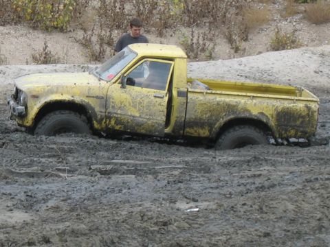1987 Ford Bronco - A yellow pickup truck is stuck in thick mud, with a person standing behind it.
