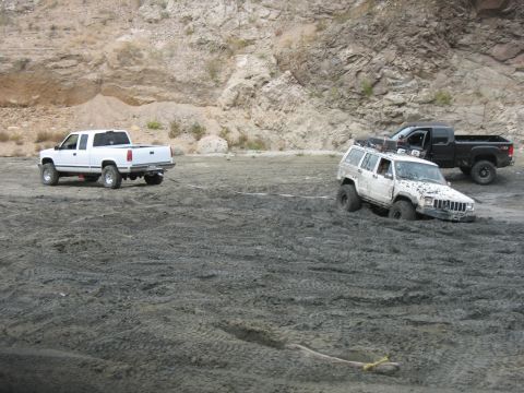 1987 Ford Bronco - A white pickup truck tows a white Jeep Cherokee that is stuck in the mud, with another black pickup truck nearby.