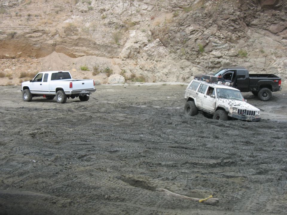 White pickup truck pulling a white Jeep Cherokee stuck in mud, with a black pickup truck behind the Jeep