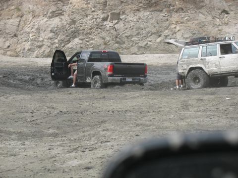 1987 Ford Bronco - Two off-road vehicles are stuck in thick mud near a rocky hillside.